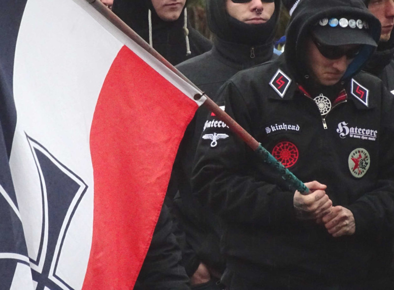 Der Teilnehmer eines neonazistischen Aufmarsches im Jahr 2016 in Magdeburg präsentiert neben zwei Schwarzen Sonnen den Reichsadler, die Reichskriegsfahne, Hatecore-Schriftzüge sowie am Kragenspiegel die germanische Jera-Rune.