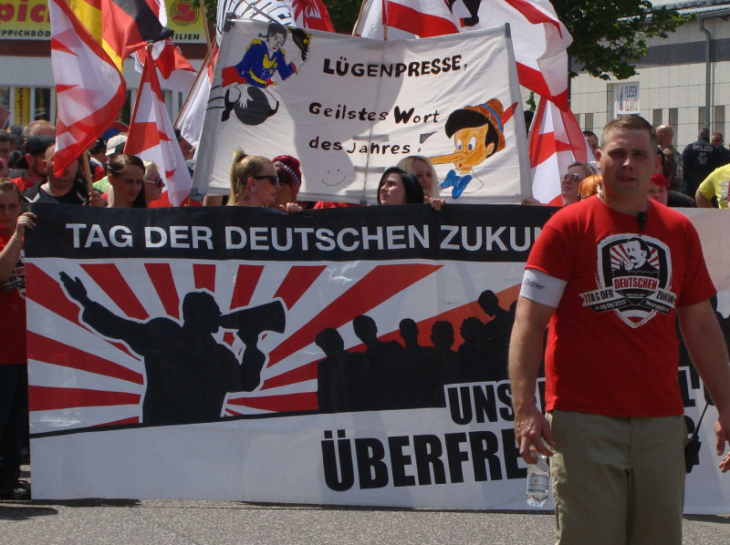 Das Wort »Lügenpresse« wurde von der Jury der Sprachkritischen Aktion zum »Unwort des Jahres 2014« gekürt. In der Erklärung der Jury heißt es: »Das Wort „Lügenpresse“ war bereits im Ersten Weltkrieg ein zentraler Kampfbegriff und diente auch den Nationalsozialisten zur pauschalen Diffamierung unabhängiger Medien. Gerade die Tatsache, dass diese sprachgeschichtliche Aufladung des Ausdrucks einem Großteil derjenigen, die ihn seit dem letzten Jahr als „besorgte Bürger“ skandieren und auf Transparenten tragen, nicht bewusst sein dürfte, macht ihn zu einem besonders perfiden Mittel derjenigen, die ihn gezielt einsetzen. Dass Mediensprache eines kritischen Blicks bedarf und nicht alles, was in der Presse steht, auch wahr ist, steht außer Zweifel. Mit dem Ausdruck „Lügenpresse“ aber werden Medien pauschal diffamiert, weil sich die große Mehrheit ihrer Vertreter bemüht, der gezielt geschürten Angst vor einer vermeintlichen „Islamisierung des Abendlandes“ eine sachliche Darstellung gesellschaftspolitischer Themen und differenzierte Sichtweisen entgegenzusetzen. Eine solche pauschale Verurteilung verhindert fundierte Medienkritik und leistet somit einen Beitrag zur Gefährdung der für die Demokratie so wichtigen Pressefreiheit, deren akute Bedrohung durch Extremismus gerade in diesen Tagen unübersehbar geworden ist«.
Das Bild zeigt den Aufmarsch am »Tag der deutschen Zukunft« am 6. Juni 2015 in Neuruppin.