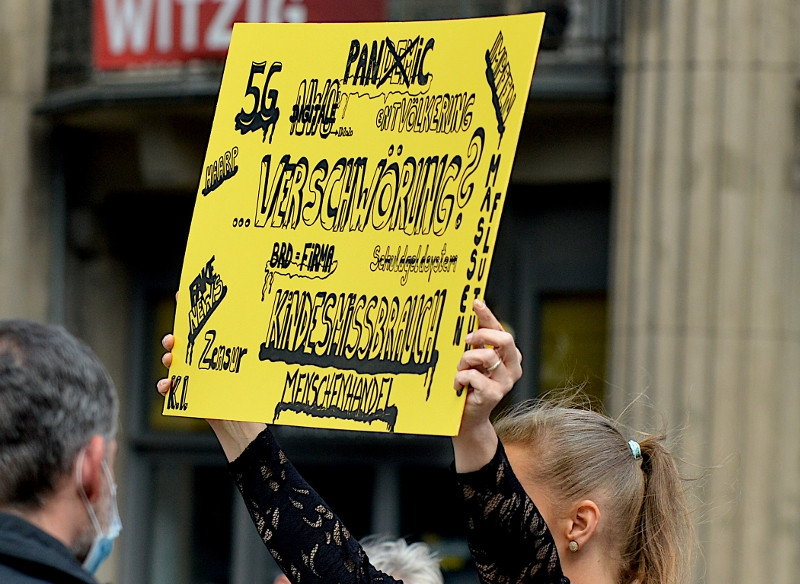 Teilnehmerin der Demonstration gegen die Maßnahmen zur Eindämmung der Corona-Pandemie am 29. August 2020 in Berlin. Auf ihrem Schild werden verschiedenen Verschwörungsmythen miteinander verbunden: Die COVID-19-Pandemie, 5G, Impfungen, Neue Weltordnung (NWO), "Entvölkerung" und "Massenflutung", HAARP und andere.
Die Wortkombination "Kindesmissbrauch Menschenhandel" spielt auf die Adrenochrom-Verschwörung an.