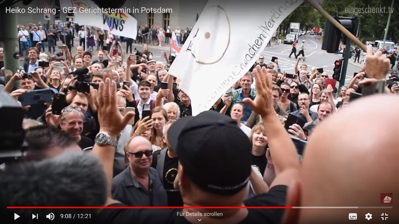 Heiko Schrang ist ein Verschwörungsideologe mit Geschäftssinn und dem Gebaren eines Rockstars. Am 10. Juli 2020 stand er wegen der Verweigerung der Zahlung der GEZ-Gebühren in Potsdam vor Gericht und ließ sich vor dem Justizgebäude von 200 angereisten Anhänger*innen mit Sprechchören feiern. Die Videoplattform Eingeschenkt TV berichtete darüber. Eingeschenkt TV ist ein Sprachrohr der selbsternannten »Corona-Rebellen« und greift in seiner Berichterstattung rechtspopulistische Themen und Argumente auf. Zu Eingeschenkt TV siehe https://www.psiram.com/de/index.php/Eingeschenkt_TV.
Screenshot: Youtube, Eingeschenkt TV, https://www.youtube.com/watch?v=3dwHPYp62HY