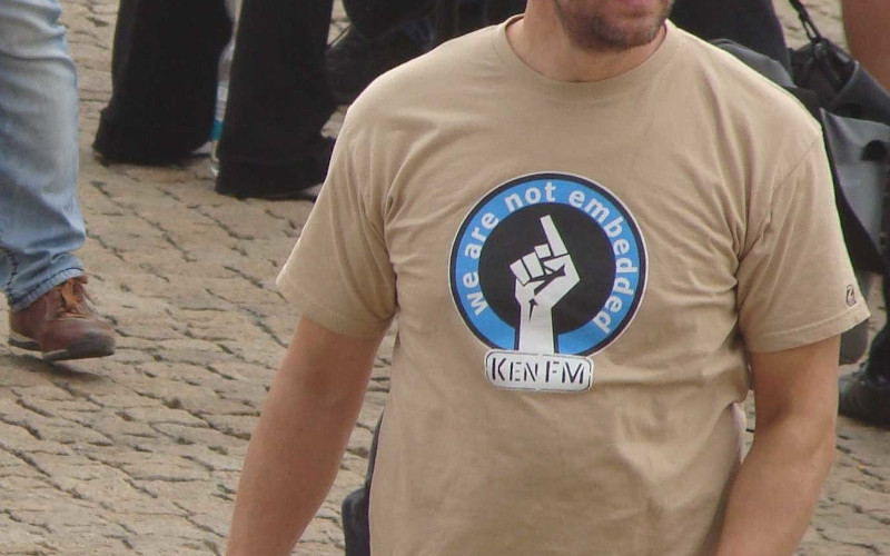 Shirt-Motiv von KenFM: »We are not embedded«. »Embedded« Journalismus beschreibt einen Journalismus, der in ein herrschaftspolitisches oder militärisches Konzept »eingebettet« ist und sich zum Werkzeug und Sprachrohr einer Regierung macht. Der Begriff geht auf den Irakkrieg im Jahr 2003 zurück. Die amerikanischen Streitkräfte verlangten von Journalist:innen, die aus den Kampfzonen berichten durften, eine Kooperation. Darüber gelang es ihnen, deren Berichterstattung maßgeblich zu beeinflussen und zu kontrollieren. »We are not embedded« meint demnach einen Journalismus, der unabhängig ist und nicht gesteuert wird.
Das Bild stammt von einer »Friedensmahnwache« in Halle an der Saale im Jahr 2014, auf dem unter anderem der rechte Verschwörungsideologe Heiko Schrang auftrat.