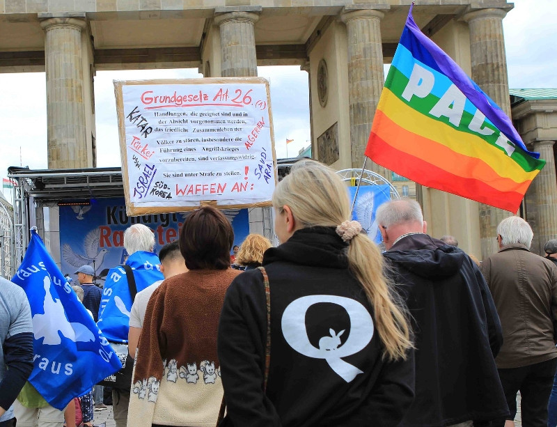 Teilnehmerin einer Kundgebung unter dem Motto »Kündigt Ramstein Air Base jetzt« am 30. Mai 2020 am Brandenburger Tor in Berlin. Die Kundgebung wurde nach Angabe der Organisator*innen von einem breiten Bündnis von Gruppierungen aus der Friedensbewegung getragen. Tatsächlich nahmen an der Kundgebung auch extreme Rechte sowie Anhänger*innen von Putin, Assad und des OAnon-Verschwörungsmythos teil.
Die QAnon-Phrasen »down the rabbit hole« und »follow the »white rabbit« sowie die Zeichnungen des weißen Kaninchen beziehen sich auf Lewis Carrolls Erzählung »Alice im Wunderland« und auf den Film »The Matrix« von 1999. In »Alice im Wunderland« folgt die Hauptfigur Alice einem weißen Kaninchen in den Bau und betritt dort eine verborgene, fantastische Welt. In »The Matrix« erhält der Protagonist Neo die Botschaft »Follow the white rabbit« und schließt sich daraufhin einer Frau an, die ein weißes Kaninchen als Tätowierung trägt. Beide verlassen ihre künstliche Scheinwelt und gelangen in die echte Welt. Das weiße Kaninchen und die sich darauf beziehenden Metaphern stehen dafür, dass Menschen beginnen, kleinen Zweifeln nachzugehen und dabei eine gigantische Verschwörung entdecken.